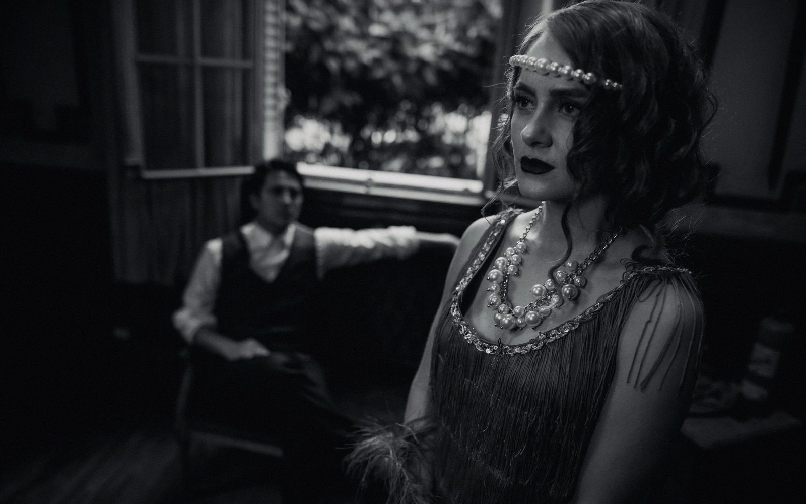 Woman in a beaded flapper dress and pearl headband stands near a seated man in vest