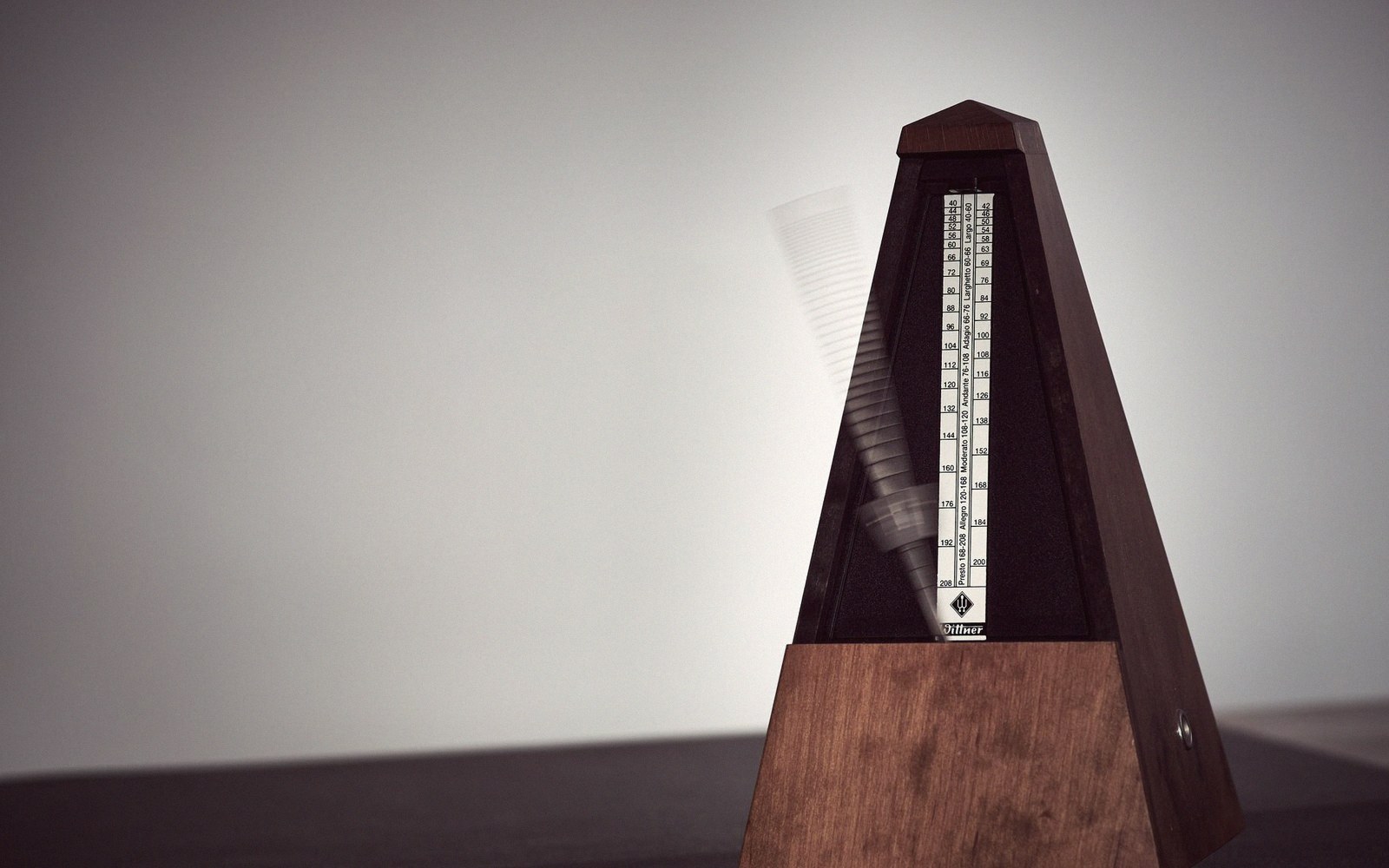Wooden pyramid metronome with its pendulum blurred mid-swing against a plain backdrop