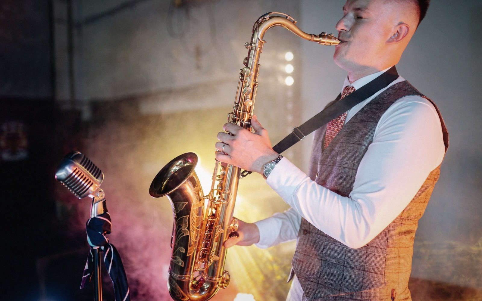 A man in a vest and tie plays saxophone on a smoky, lit stage beside a vintage microphone